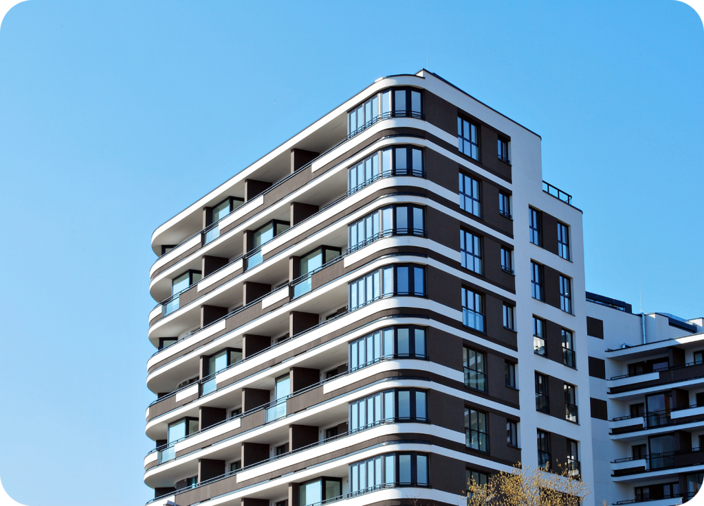 
Um prédio residencial de estilo moderno e fachada branca e marrom. A foto é tirada de baixo para cima, mostrando a arquitetura de varandas e janelas em curvas, sob um céu azul claro.