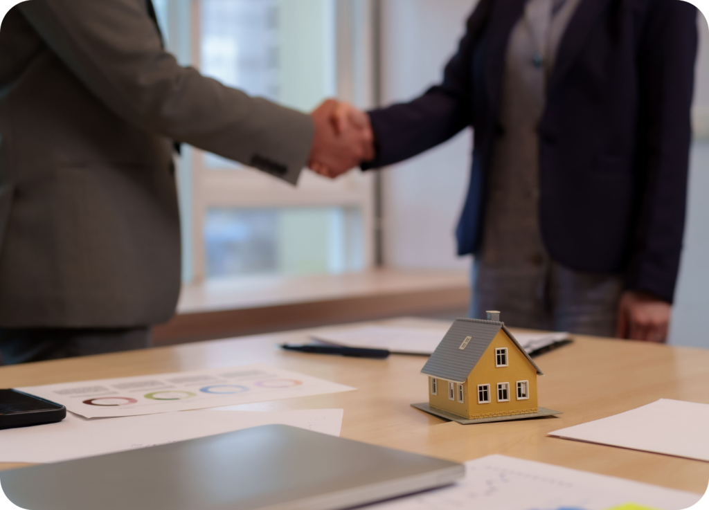 
Um close de um aperto de mãos entre duas pessoas. Uma delas veste um terno cinza, e a outra, um blazer azul-marinho. Em primeiro plano, sobre uma mesa, há papéis, um laptop fechado e um modelo em miniatura de uma casa.