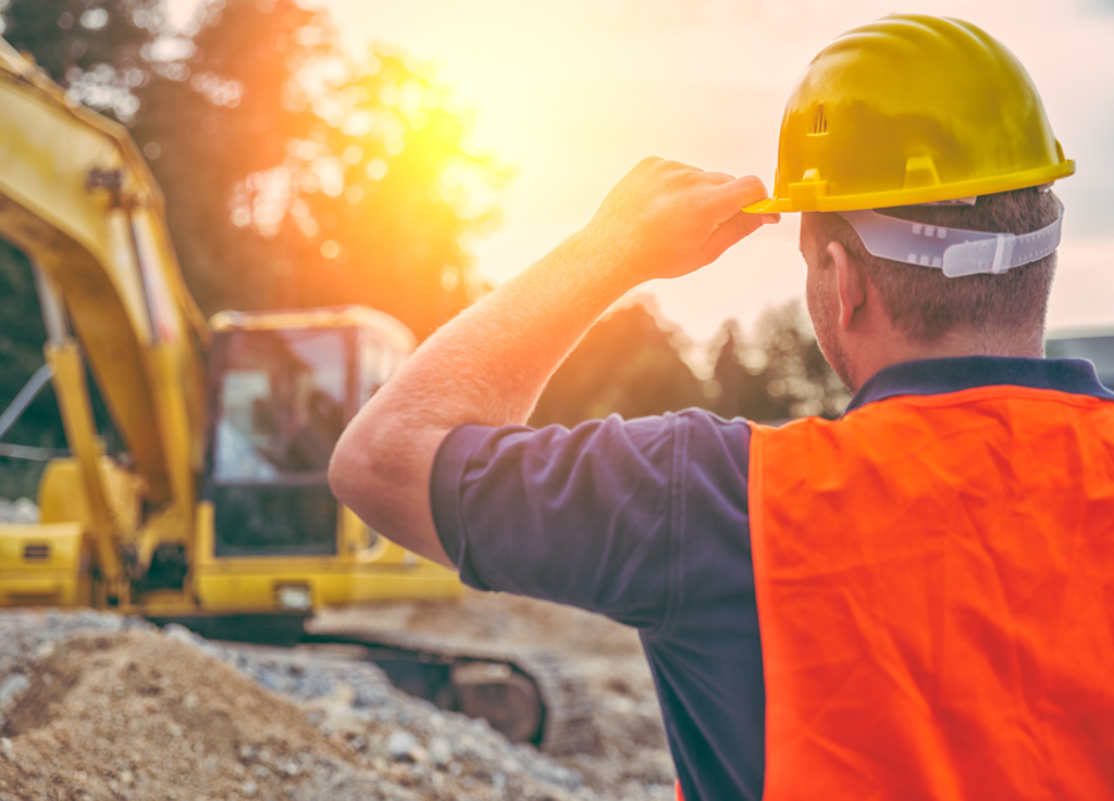Atrás, um homem de costas, vestindo uma camisa de polo azul-escura e um colete laranja de alta visibilidade, está ajustando seu capacete de segurança amarelo. Ele está em um local de construção com montes de terra e cascalho. Ao fundo, um trator amarelo está estacionado. A luz do sol brilha intensamente, criando um forte clarão.