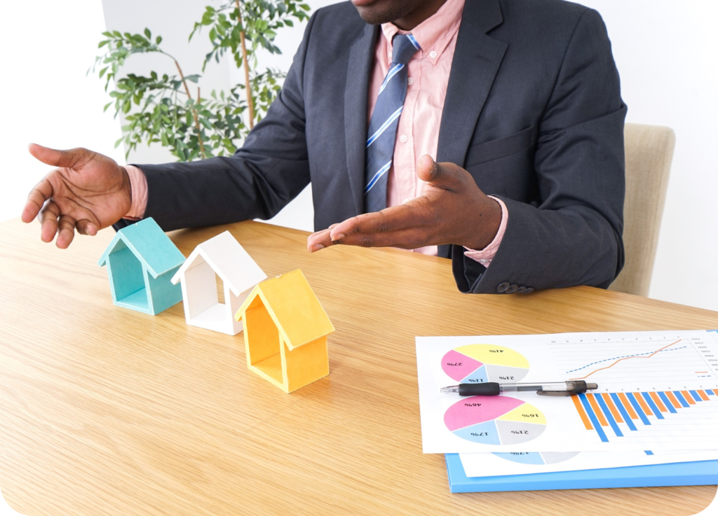 
Um homem, de terno e gravata, está sentado à mesa e usa as mãos para gesticular. Na mesa, há três modelos em miniatura de casas nas cores azul-claro, branco e amarelo. Há também papéis com gráficos de pizza e de barras, além de uma caneta.