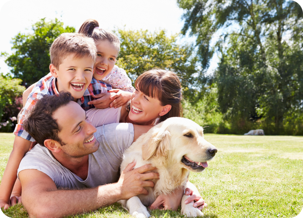 Uma família, composta por um casal e duas crianças, está no gramado de um jardim ensolarado. Eles estão deitados no chão, abraçando um grande cachorro. A família sorri para a câmera, com a garota e o menino sobre as costas do pai, e a mãe segurando o cachorro. O cachorro parece ser um golden retriever.