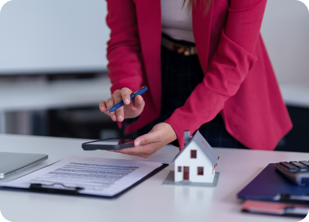 
Uma pessoa vestindo um blazer rosa-choque está usando uma caneta para assinar um documento em um tablet. Na mesa, há um modelo em miniatura de uma casa, papéis em uma prancheta e um laptop. A imagem está com foco nas mãos da pessoa e no tablet.