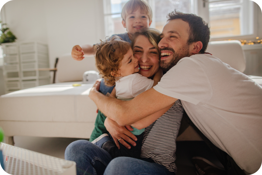 Família feliz e sorridente (pai, mãe e dois filhos pequenos) abraçada em um apartamento moderno, com luz natural abundante, transmitindo a sensação de conforto, segurança e viver bem. A imagem reforça o valor de morar em um empreendimento de alto padrão no Vieiralves.