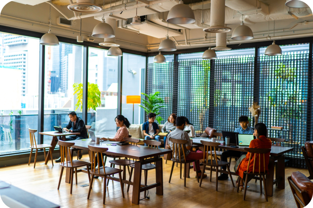 Espaço de coworking aconchegante em condomínio com grandes janelas e vista para a cidade. O ambiente possui diversas luminárias pendentes, mesas de madeira escura com cadeiras e poltronas, onde várias pessoas trabalham em laptops ou conversam em áreas de convivência.