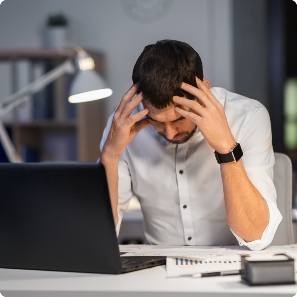 Um homem jovem, vestindo uma camisa social branca e relógio de pulso, está sentado à frente de um notebook em um ambiente de escritório com iluminação suave. Ele está com a cabeça baixa e as mãos apoiadas nas têmporas, demonstrando cansaço, estresse ou preocupação ao analisar documentos e gráficos espalhados sobre a mesa, representando as dificuldades ou desafios em processos burocráticos e financeiros.