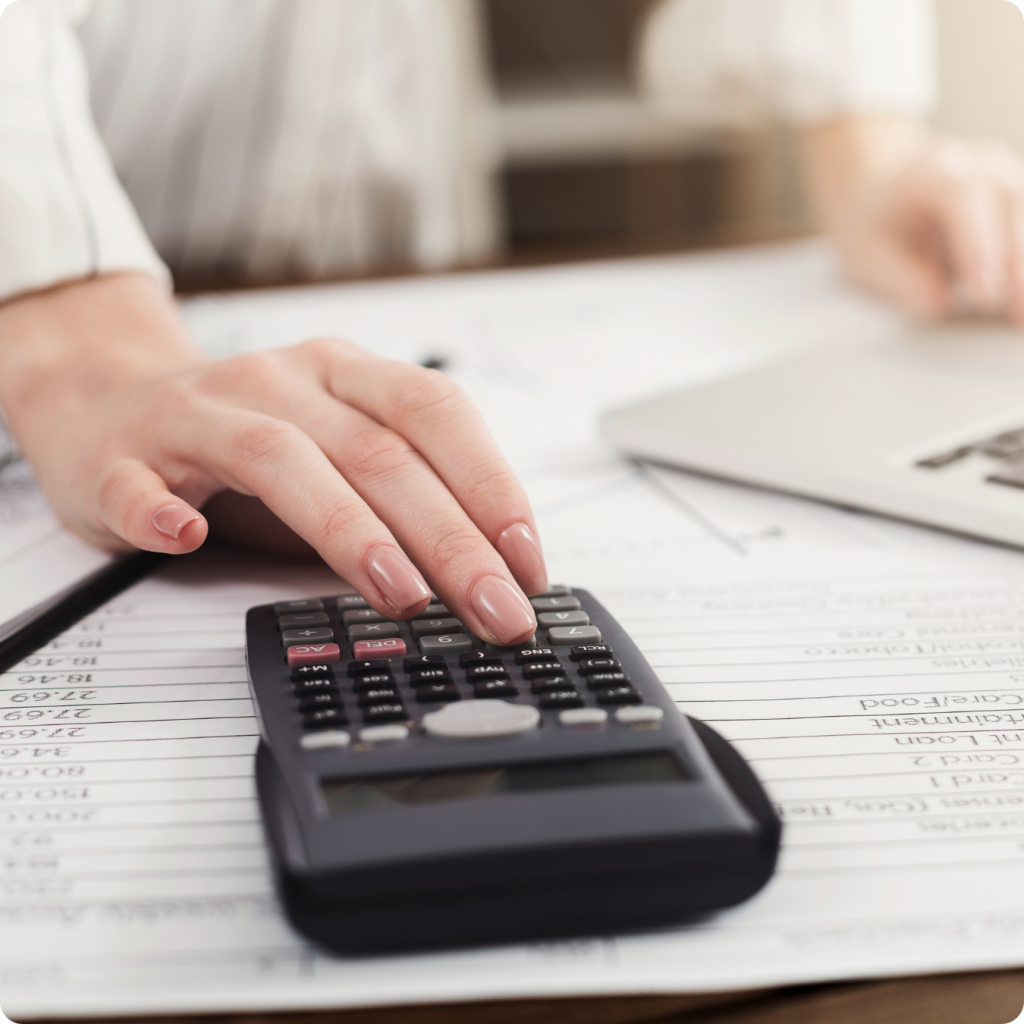 Uma mão feminina com unhas pintadas de nude utiliza uma calculadora financeira preta sobre uma mesa repleta de planilhas e documentos com números. Ao fundo, de forma desfocada, é possível ver parte de um notebook e a pessoa concentrada no trabalho, transmitindo uma ideia de planejamento, cálculo de rendimentos e análise financeira detalhada.